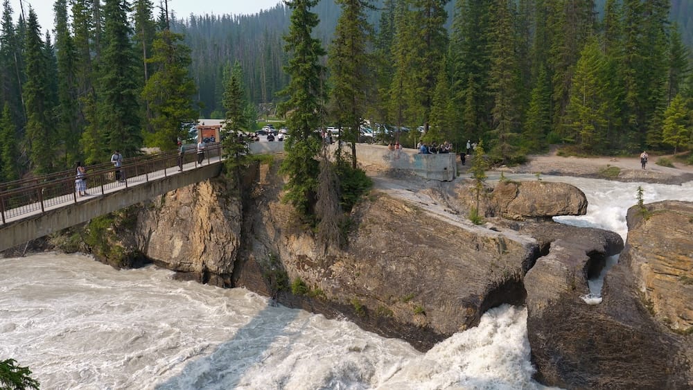Natural Bridge and Kicking Horse River viewpoint in Yoho National Park, British Columbia, Canada Natural Bridge in Yoho National Park, British Columbia, Canada, with the Kicking Horse River rushing through narrow rock channels beside a viewing platform, forested slopes and several visitors.