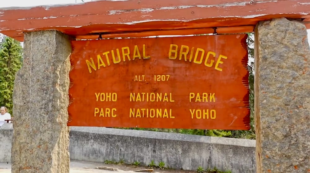 Orange Natural Bridge sign mounted between stone posts at the visitor area in Yoho National Park, British Columbia, Canada, with bilingual park lettering and evergreen forest behind it.