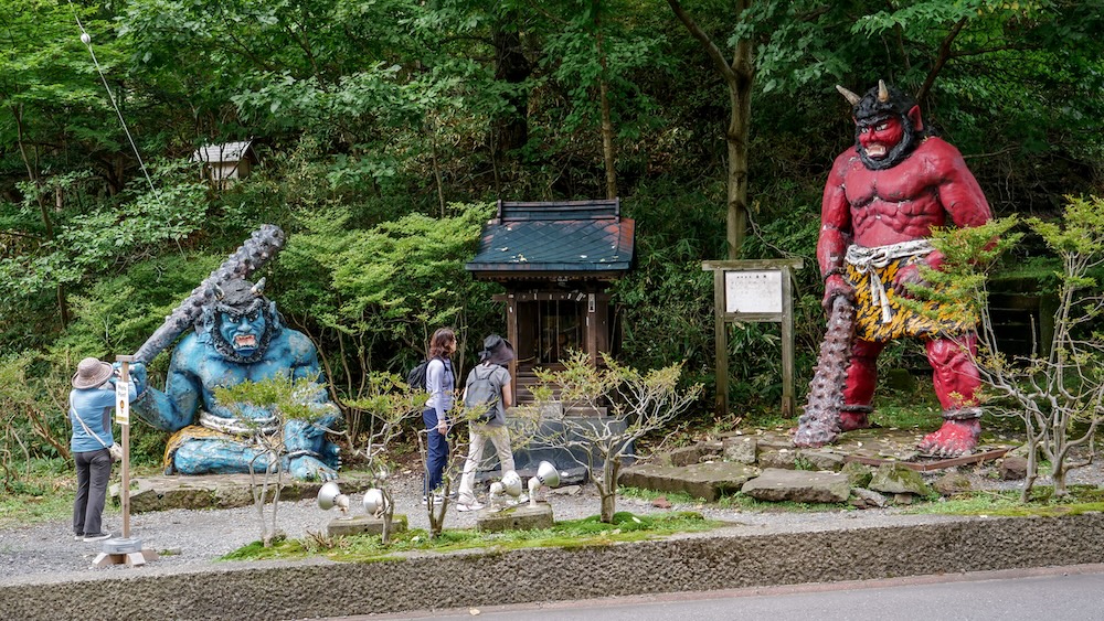 Guardianes oni rojo y azul del Santuario Nembutsu cerca de Yuzawa Shrine, Noboribetsu Onsen, Hokkaido, Japón Estatuas de un oni rojo de pie y un oni azul sentado, ambos con grandes garrotes de púas, junto a un pequeño santuario de madera cerca de Yuzawa Shrine, en Noboribetsu Onsen, Hokkaido, Japón, mientras varios turistas recorren el lugar y sacan fotografías.