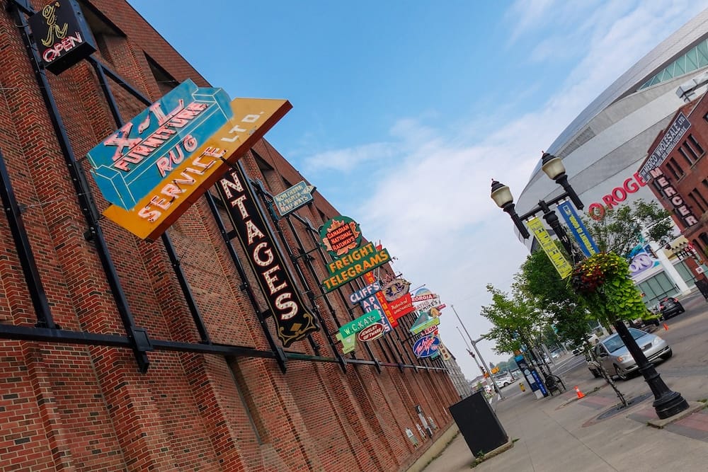 Historic commercial signs line a brick wall at the Neon Sign Museum in downtown Edmonton, Alberta, Canada, with Rogers Place, street lamps and blue sky visible along the city streetscape.