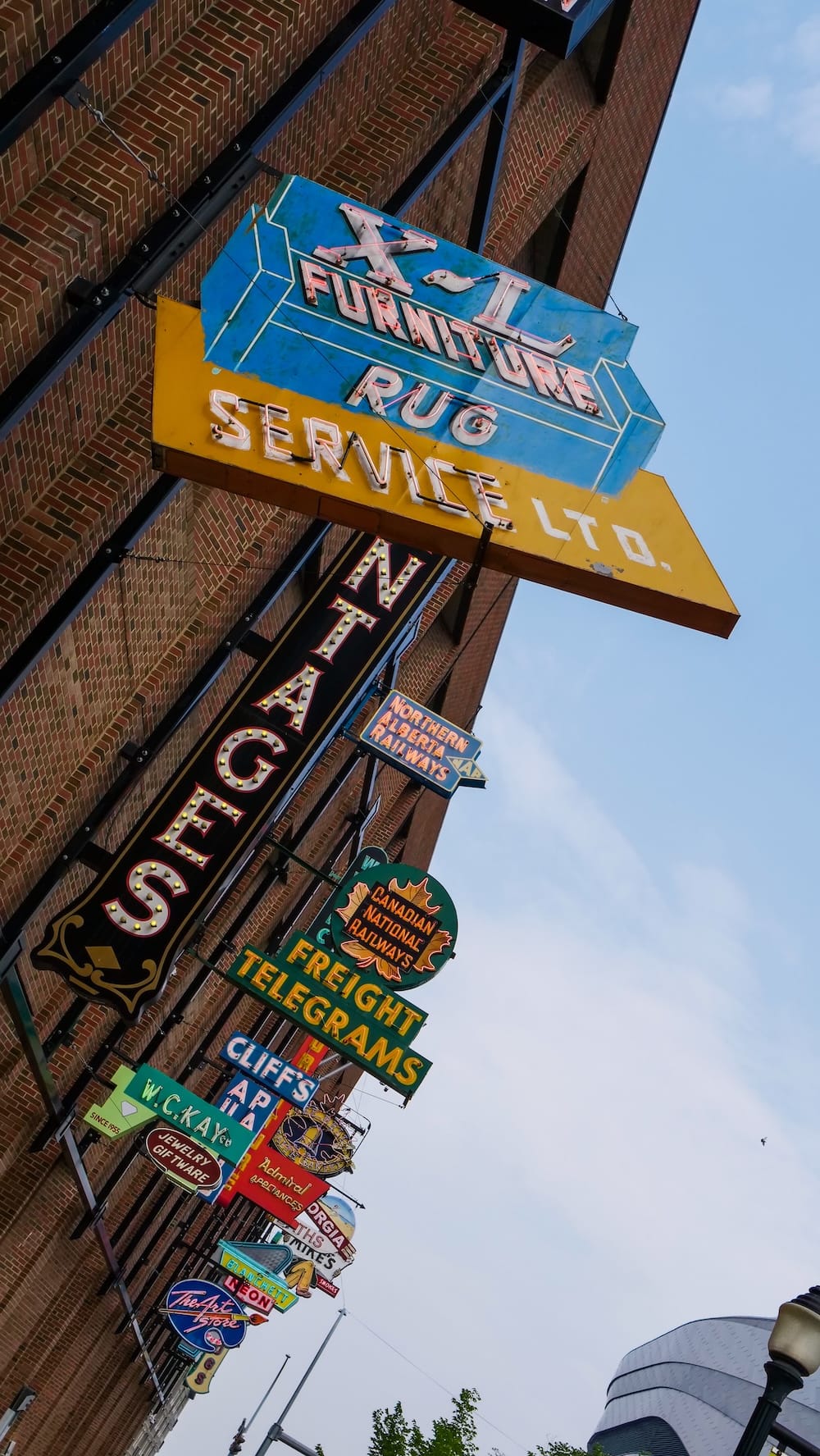 Hilera vertical de carteles de neón en el Neon Sign Museum, 104 Street, Edmonton, Alberta, Canadá Una hilera vertical de carteles de neón en un edificio de ladrillo, encabezada por X-L Furniture Rug Service y Pantages, bajando junto a Northern Alberta Railways, Canadian National Railways y Cliff's Auto Parts, con Rogers Place visible más abajo en el centro de Edmonton, Alberta, Canadá.