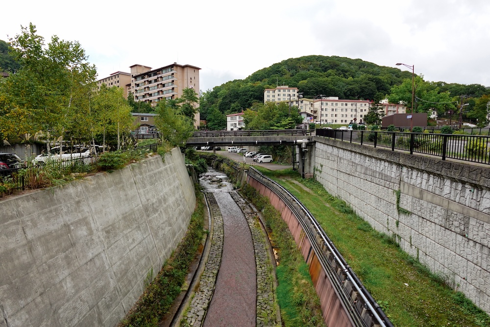 Vista de Noboribetsu Onsen, en Hokkaido, Japón, con un canal de drenaje revestido de hormigón que pasa bajo un puente, hoteles termales de varios pisos, árboles junto a la calle y colinas cubiertas de bosque al fondo.
