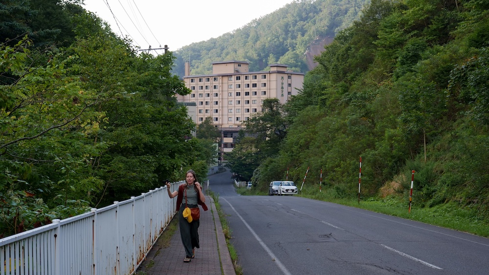 Morning roadside walk toward the Oyunuma River Natural Footbath, Noboribetsu Onsen, Hokkaido, Japan – Audrey Bergner Audrey Bergner walks along a narrow paved footpath beside a quiet road and a white railing, with forested hills and a large hotel ahead, in Noboribetsu Onsen, Hokkaido, Japan.