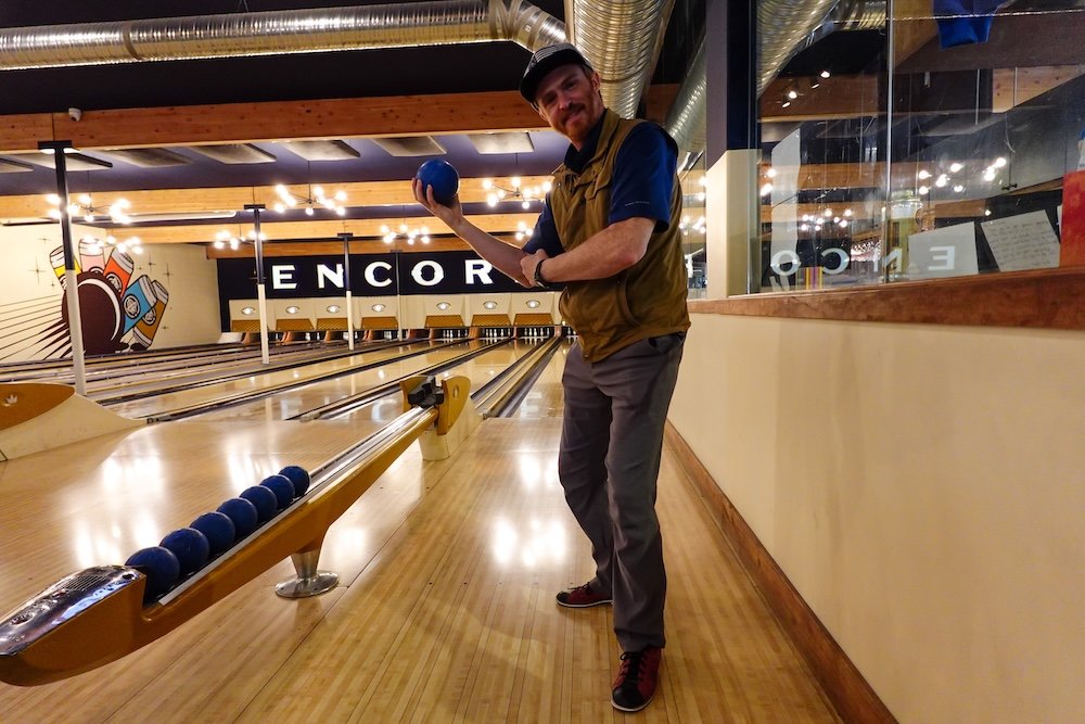Nomadic Samuel bowling at Encore Brewing Co. Nomadic Samuel Jeffery poses mid-throw with a blue bowling ball at Encore Brewing Co. in Cranbrook, standing beside the lane’s return rack with rows of balls visible and the lively, neon-lit bowling alley stretching out behind him.