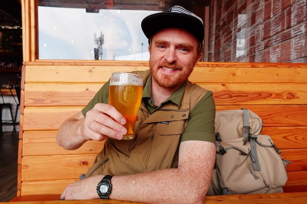 Nomadic Samuel enjoying Fernie Brewing pint in Cranbrook Nomadic Samuel Jeffery raises a cold pint of Fernie Brewing Company beer while relaxing in a wooden booth at Fire Hall Kitchen & Tap, Cranbrook’s heritage gastropub known for craft drinks and casual bites, enjoying a refreshing break after sightseeing.