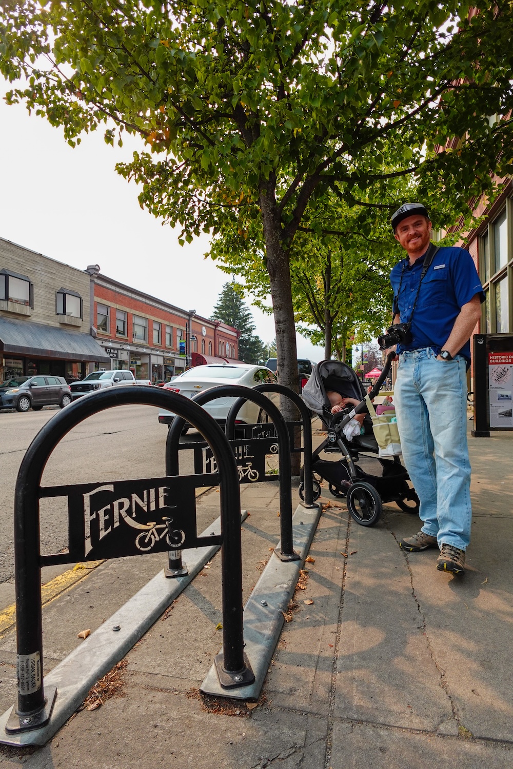 Fernie Heritage Walk after visiting the Fernie Museum Nomadic Samuel walking Fernie’s Heritage Walk after visiting the Fernie Museum, pushing a stroller with baby Aurelia and holding a camera along historic 2nd Avenue, where brick heritage buildings, bike racks, and leafy sidewalks define downtown Fernie.