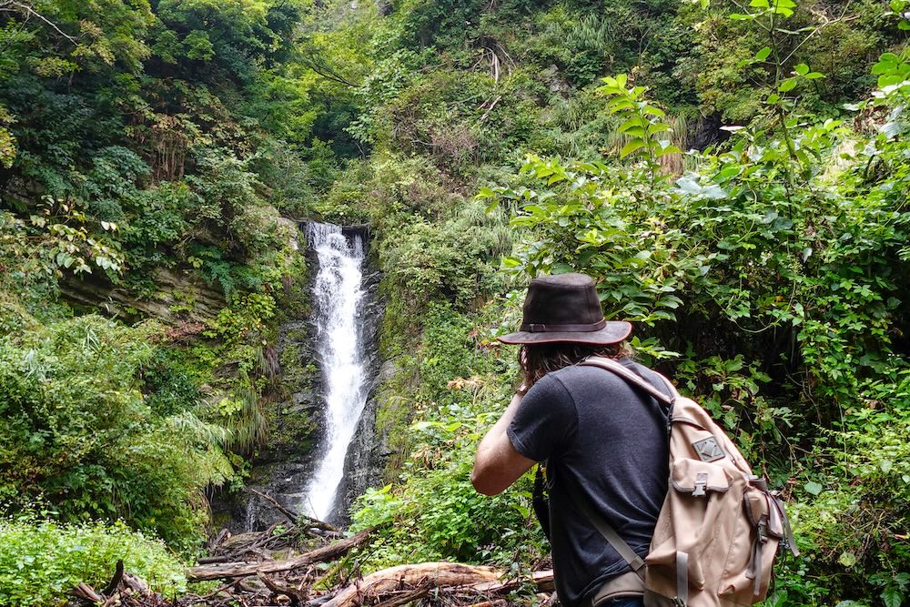 Here I Am Nomadic Samuel taking photos of r scenic waterfall in Japan in the lush countryside 