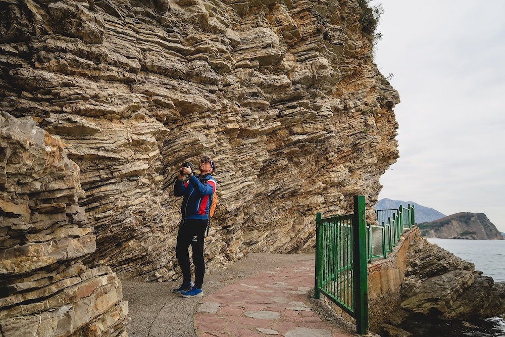 Nomadic Samuel taking photos of the jagged coastline on a walk in Budva, Montenegro 