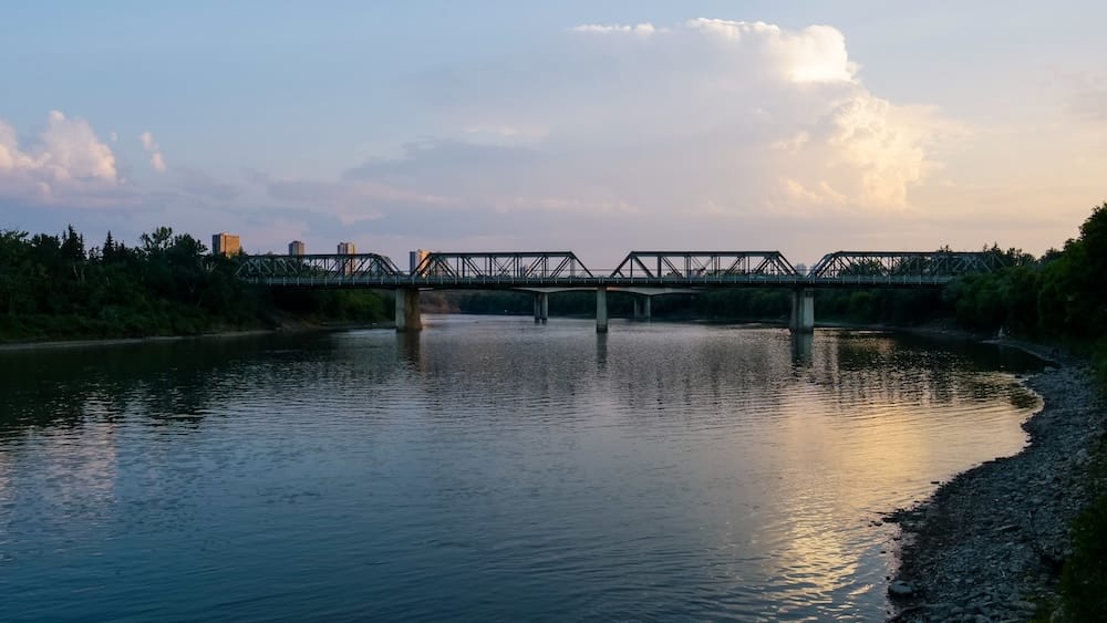 Puente del Valle del Río North Saskatchewan al Atardecer, Edmonton, Alberta, Canadá Un puente de estructura metálica cruza el tranquilo río North Saskatchewan al atardecer en Edmonton, Alberta, Canadá, con una luz cálida reflejada en el agua y el perfil del centro visible a lo lejos.