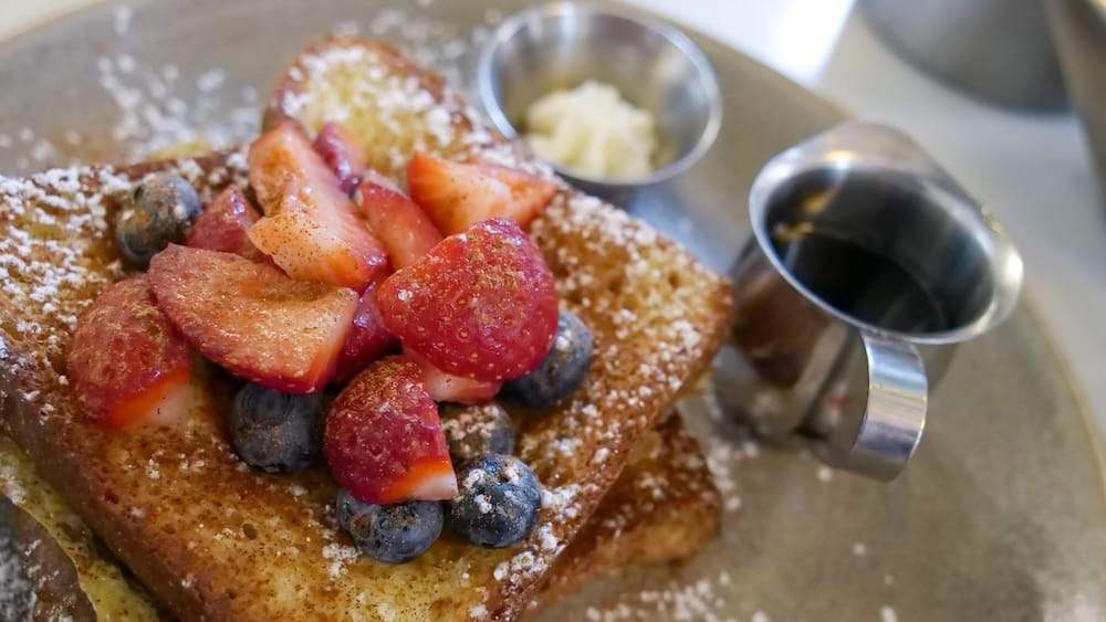 Primer plano de unas tostadas francesas con canela en OEB Breakfast Co. cubiertas con frutillas, arándanos y azúcar impalpable, con un acompañamiento de jarabe de arce y manteca en un plato gris piedra.