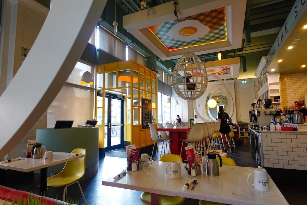 El luminoso interior de OEB Breakfast Co. en el centro de Edmonton, con mesas con tapa de mármol, sillas amarillas, jarras de café y un mural de huevos y estampado a cuadros en el techo sobre el salón comedor.