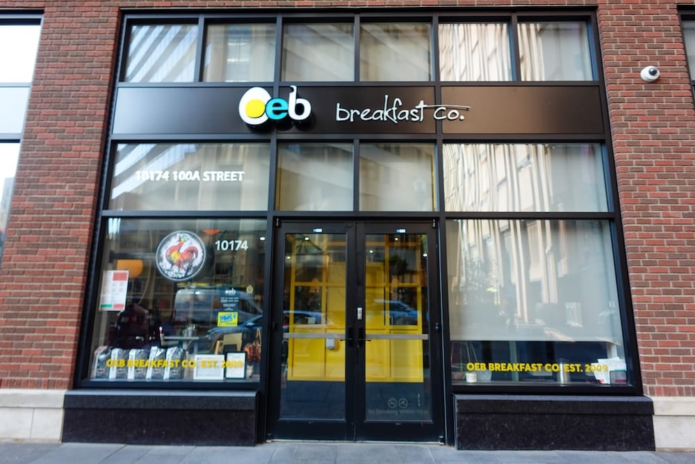 La fachada de ladrillo y marcos negros de OEB Breakfast Co. en 10174 100A Street en el centro de Edmonton, Alberta, con el logo de "Est. 2009" y las puertas de entrada amarillas a la vista.