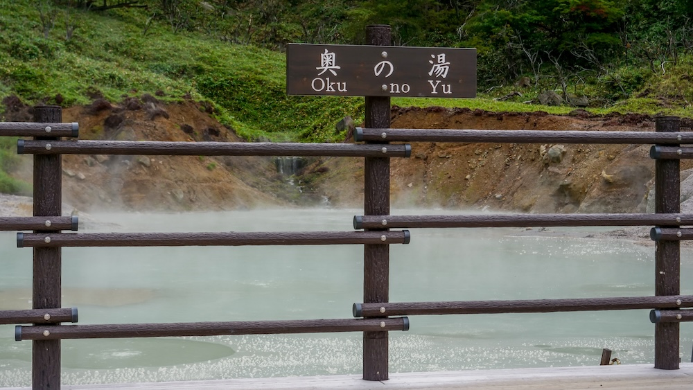 Oku No Yu Geothermal Pool in Noboribetsu Onsen, Hokkaido, Japan – Steaming Water and Wooden Viewing Platform Steaming milky-blue geothermal water at Oku No Yu in Noboribetsu Onsen, Hokkaido, Japan, viewed behind a wooden safety railing with a Japanese and English sign, exposed mineral-stained earth and green forest vegetation.