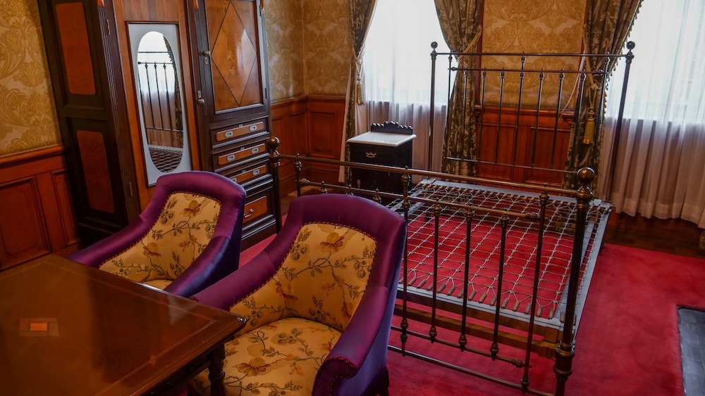 Historic Bedroom and Period Furniture Inside the Old Public Hall of Hakodate Ward in Hokkaido, Japan Historic bedroom inside the Old Public Hall of Hakodate Ward in Hakodate, Hokkaido, Japan, featuring an antique iron bed, two purple upholstered armchairs, a wooden mirrored wardrobe and ornate patterned wallpaper.