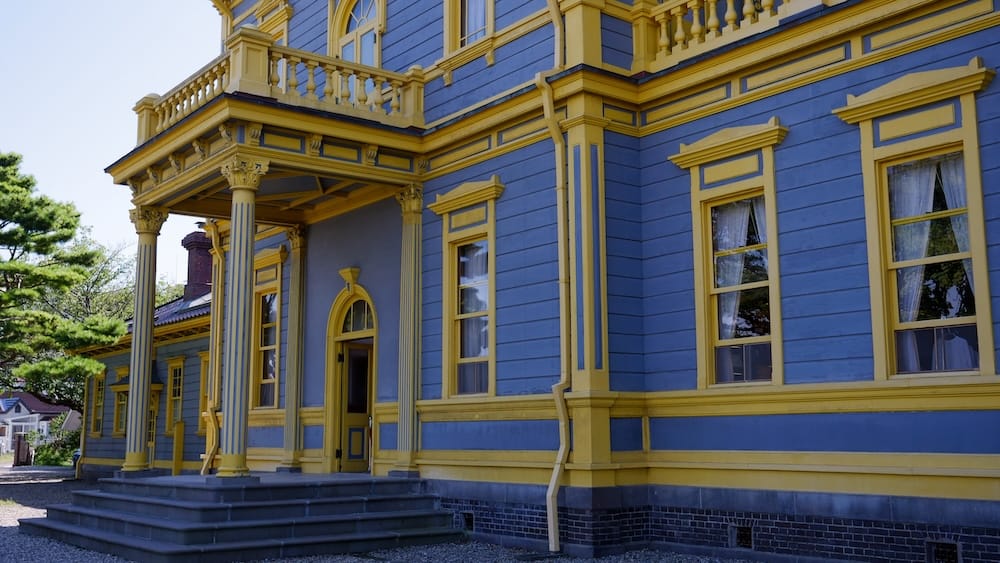 A side view of the Old Public Hall of Hakodate Ward in Hakodate, Hokkaido, Japan, showing its blue wooden clapboard walls, yellow trim, fluted columns, balcony and stone entrance steps on a clear day.