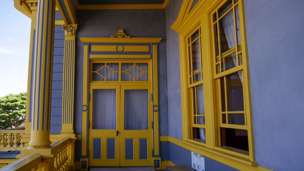 Yellow and blue-grey double doors, diamond-pane transom windows, and a fluted column topped with carved acanthus leaves on the second-floor balcony of the Old Public Hall of Hakodate Ward in Motomachi, Hakodate, Hokkaido, Japan.