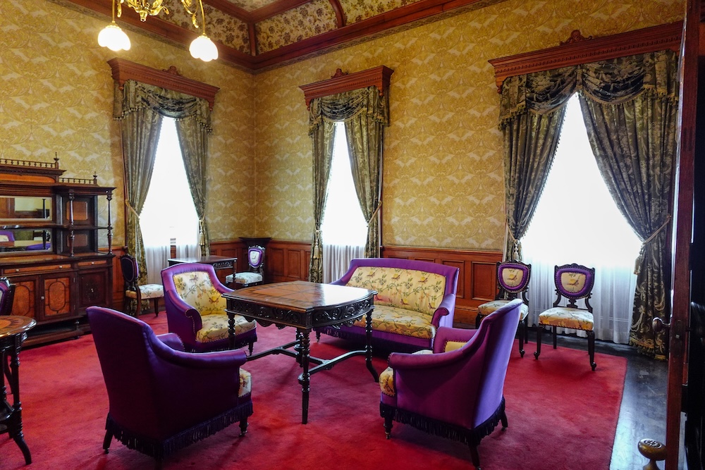 Restored upstairs Imperial sitting room inside the Old Public Hall of Hakodate Ward in Hakodate, Hokkaido, Japan, featuring purple velvet-trimmed armchairs, a carved wooden table, patterned gold wallpaper, and heavy drapes over red carpet.