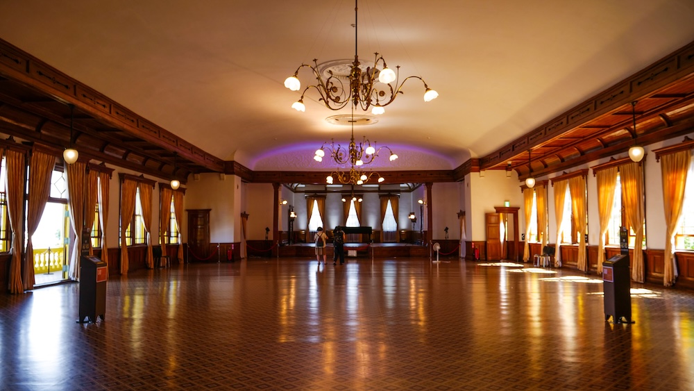 Wide view of the six-metre-high Great Hall on the second floor of the Old Public Hall of Hakodate Ward in Hakodate, Hokkaido, Japan, showing polished parquet flooring, ornate brass chandeliers, tall draped windows, and the stage.