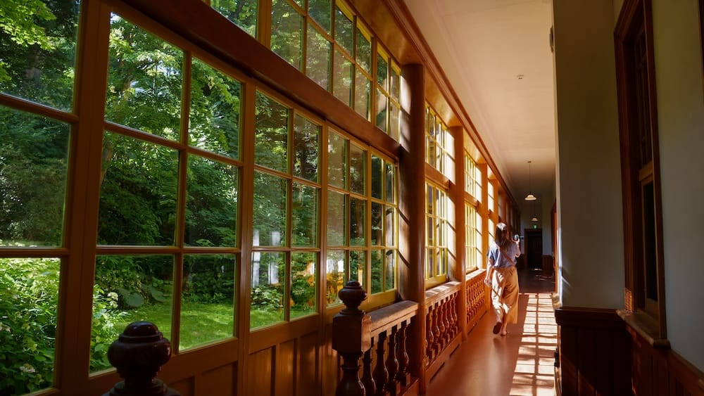 Audrey Bergner walks through a sunlit corridor inside the Old Public Hall of Hakodate Ward in Hakodate, Hokkaido, Japan, beside tall wooden-framed windows overlooking the green garden outside.