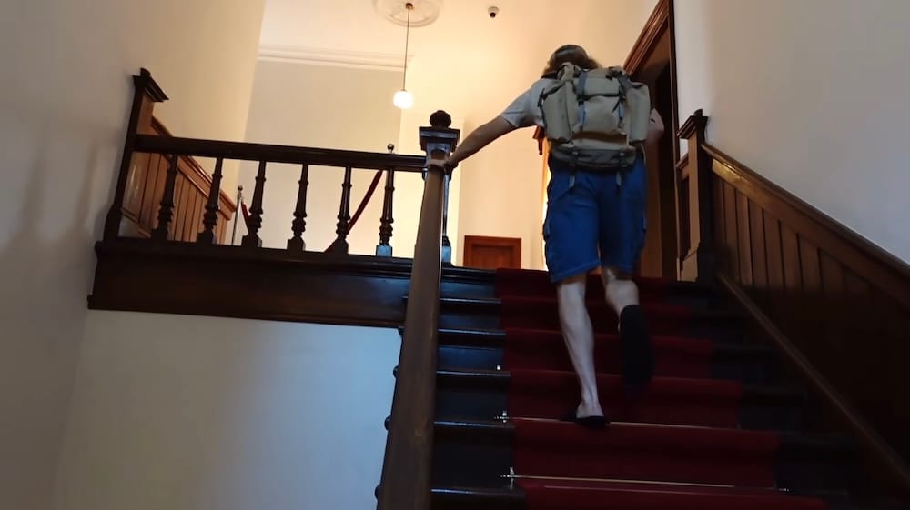 Interior staircase at the Old Public Hall of Hakodate Ward, Hakodate, Hokkaido, Japan – Samuel Jeffery Samuel Jeffery climbs a red-carpeted wooden staircase inside the Old Public Hall of Hakodate Ward in Hakodate, Hokkaido, Japan, wearing a backpack and holding the dark polished handrail.
