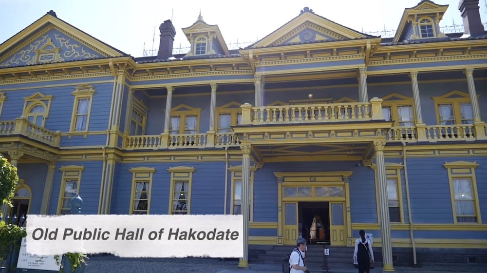 The blue and yellow two-storey facade of the Old Public Hall of Hakodate Ward in Motomachi, Hakodate, Hokkaido, Japan, with a columned balcony, dormer windows and visitors at the entrance under a clear sky.