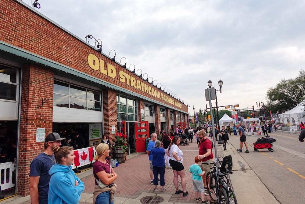 Visitors gather outside the brick exterior of Old Strathcona Farmers' Market, its yellow signage lit above roll-up doors, along Whyte Avenue in Edmonton, Alberta, Canada, with string lights, a Canadian flag, and street tents visible down the block.