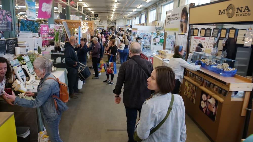 Audrey Bergner walking through the busy indoor Old Strathcona Farmers’ Market in Edmonton, Alberta, Canada, with shoppers, food stalls and vendor displays filling the aisles.