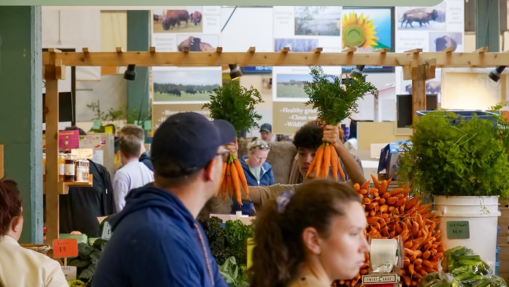A vendor holds up bunches of fresh carrots with their greens still attached at a produce stall inside Old Strathcona Farmers' Market in Edmonton, Alberta, Canada, with buckets of carrots and bison-farm signage in the background.