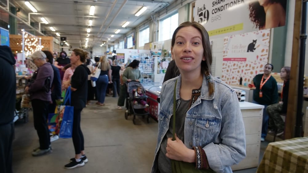 Audrey Bergner stands inside a busy indoor market in Old Strathcona, Edmonton, Alberta, Canada, surrounded by vendor stalls, shoppers and local food and craft displays.