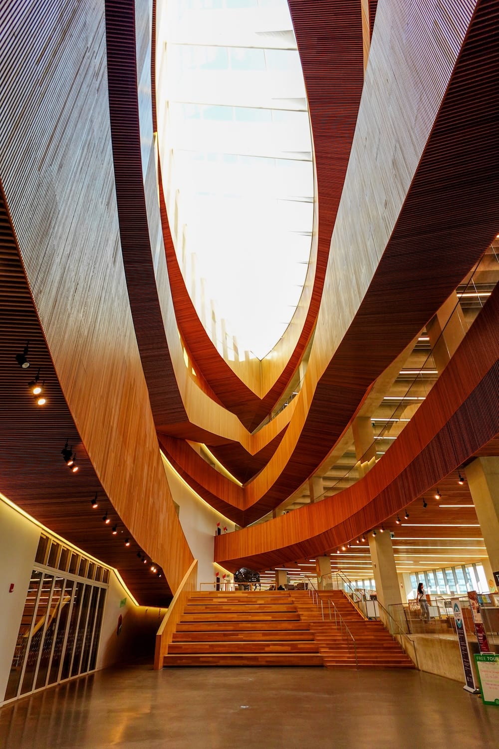 El atrio de madera ovalado con forma de canoa de la Biblioteca Central de Calgary en Calgary, Alberta, Canadá, mirando hacia un tragaluz, con niveles de balcones curvos y una amplia escalera central abajo.