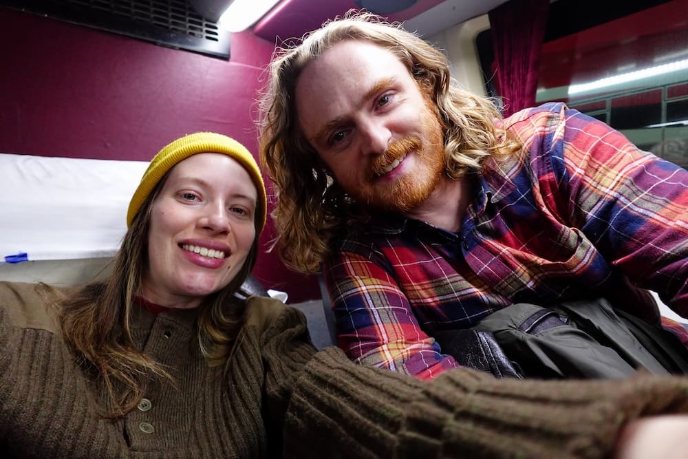 Samuel Jeffery and Audrey Bergner Inside an Overnight Bus in Patagonia, Argentina Samuel Jeffery and Audrey Bergner smile inside a long-distance overnight bus in Patagonia, Argentina, seated beneath bright cabin lights with curtains, bunks and travel bags visible around them.