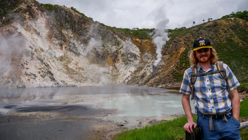 Samuel Jeffery, wearing a backpack and cap, smiles beside Oyunuma, a grey, steaming crater lake with milky water, pale volcanic cliffs and a rising plume of steam in Noboribetsu Onsen, Hokkaido, Japan.