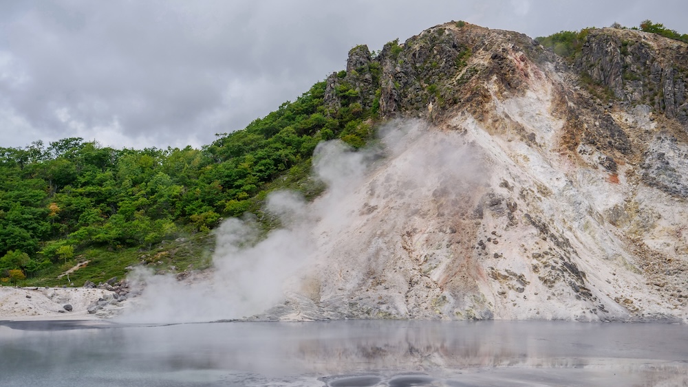 Vapor en la laguna de cráter Oyunuma y ladera volcánica en el paseo geotérmico, Noboribetsu Onsen, Japón El vapor sube por la orilla de la laguna de cráter Oyunuma bajo una ladera volcánica blanqueada y color óxido y un cerro cubierto de bosque verde bajo el cielo gris en Noboribetsu Onsen, Hokkaido, Japón.
