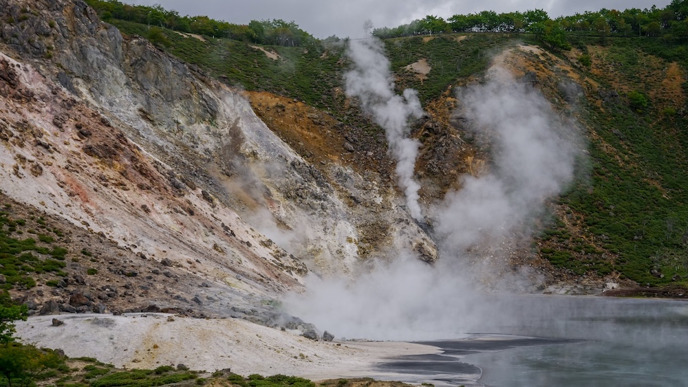 Densas columnas de vapor blanco saliendo de las fumarolas junto a Oyunuma, en Noboribetsu Onsen, Hokkaido, Japón, con agua gris azulada, laderas volcánicas claras teñidas de minerales, rocas expuestas y vegetación verde en los cerros.