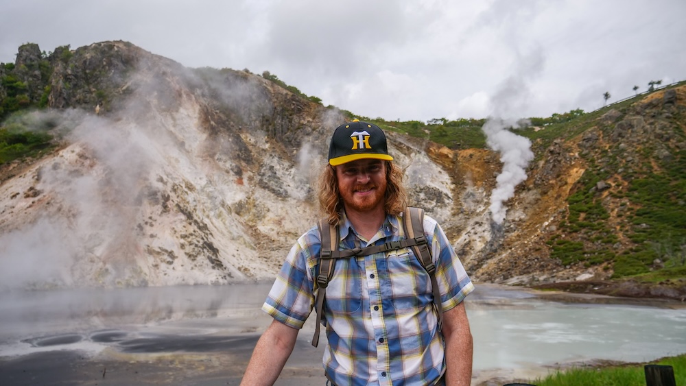 Samuel Jeffery standing beside the steaming, pale blue-grey waters of Oyunuma geothermal pond in Noboribetsu Onsen, Hokkaido, Japan, wearing a plaid shirt, baseball cap and backpack, with volcanic slopes and rising steam behind him.
