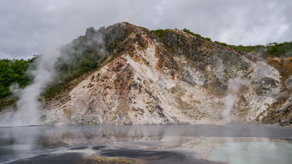 Laguna geotérmica Oyunuma cerca de Noboribetsu Onsen, Hokkaido, Japón: agua humeante y laderas volcánicas teñidas por minerales Laguna geotérmica Oyunuma humeante cerca de Noboribetsu Onsen, Hokkaido, Japón, con agua gris azulada, dibujos minerales oscuros en la orilla, vapor blanco saliendo de fumarolas y una ladera rocosa bajo un cielo nublado.
