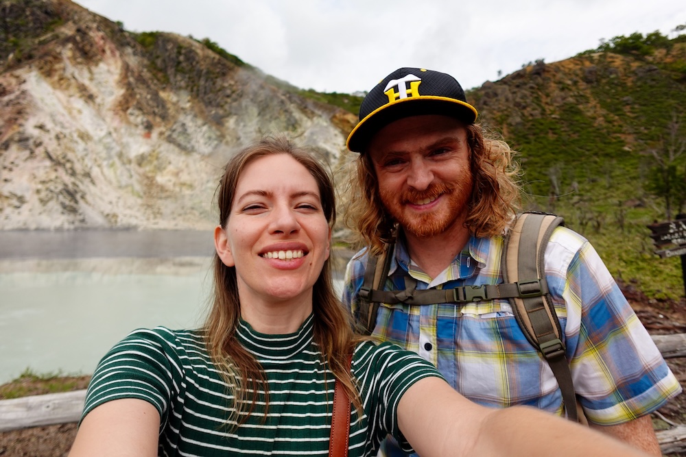 Samuel Jeffery y Audrey Bergner junto a una laguna geotérmica en Noboribetsu Onsen, Hokkaido, Japón Samuel Jeffery y Audrey Bergner sonríen mientras se sacan una selfie junto a una laguna geotérmica de color turquesa pálido en Noboribetsu Onsen, Hokkaido, Japón, con laderas volcánicas teñidas por minerales y vegetación verde detrás.