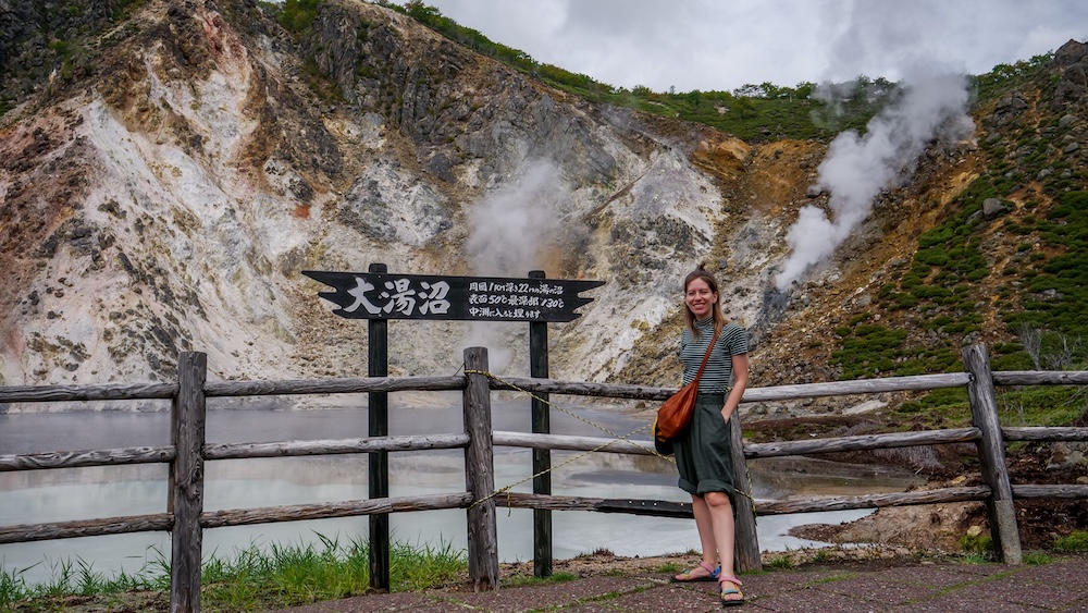 Audrey Bergner at Oyunuma Pond (大湯沼) – Japanese Landmark Sign and Steaming Volcanic Slopes, Hokkaido, Japan Audrey Bergner standing beside a wooden sign reading Oyunuma (大湯沼) at Oyunuma Pond in Noboribetsu Onsen, Hokkaido, Japan, with milky geothermal water, mineral-stained volcanic slopes and thick plumes of steam behind her.