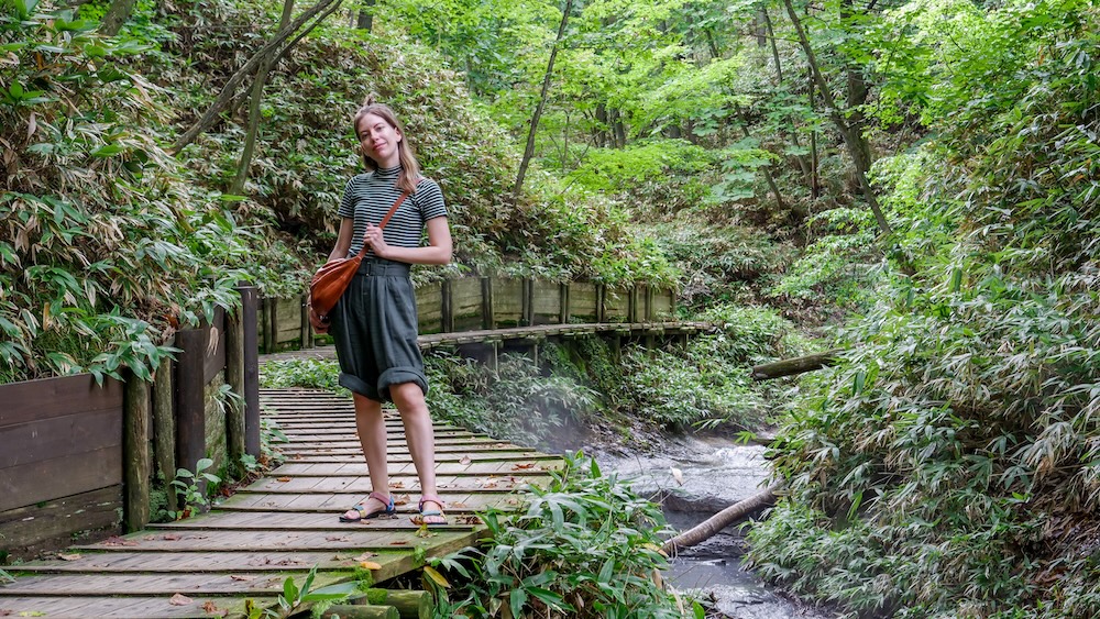 Pasarela de madera junto al humeante río Oyunuma cerca del baño de pies natural, Noboribetsu Onsen, Japón – Audrey Bergner Audrey Bergner parada en sandalias sobre una pasarela de madera junto a un arroyo humeante y cañas de bambú en el bosque cerca del baño de pies natural del río Oyunuma en Noboribetsu Onsen, Hokkaido, Japón.
