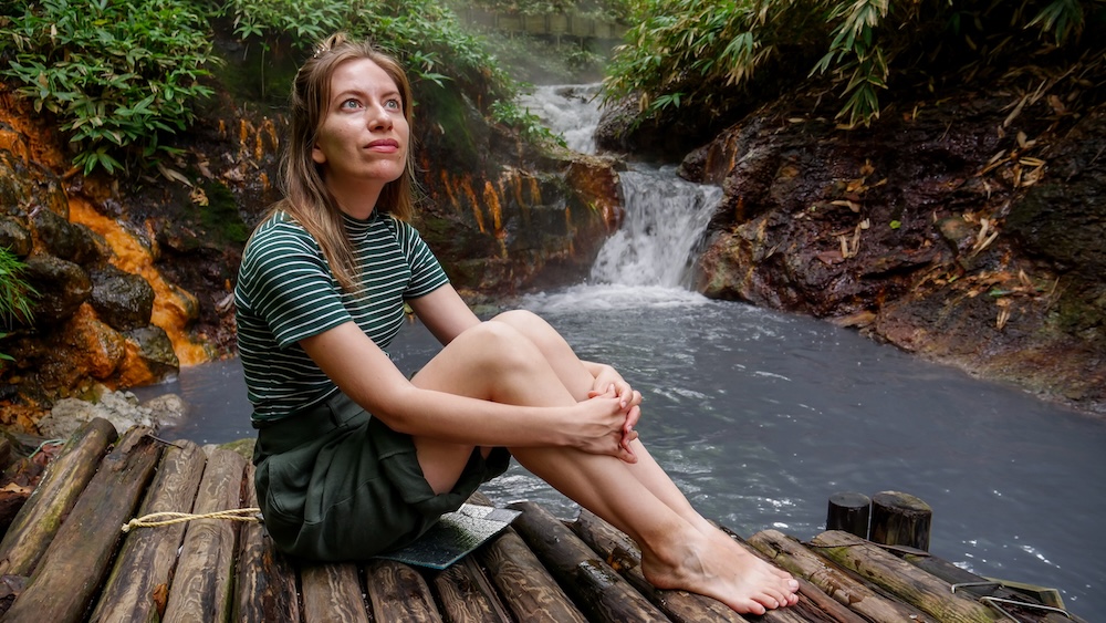 Audrey Bergner at Oyunuma River Natural Footbath, Noboribetsu Onsen, Hokkaido, Japan – Riverside Waterfall Audrey Bergner sits barefoot on a bamboo platform beside the milky-blue Oyunuma River Natural Footbath in Noboribetsu Onsen, Hokkaido, Japan, with a small waterfall, mineral-stained rocks and dense greenery behind her.