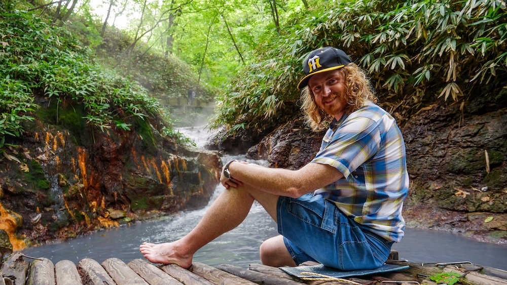 Barefoot at the Oyunuma River Natural Footbath on our walking loop, Noboribetsu Onsen, Hokkaido, Japan – Samuel Jeffery Barefoot Samuel Jeffery sits on bamboo seating and a padded mat beside the milky stream and small waterfall of the Oyunuma River Natural Footbath, with orange-stained rocks, in Noboribetsu Onsen, Hokkaido, Japan.