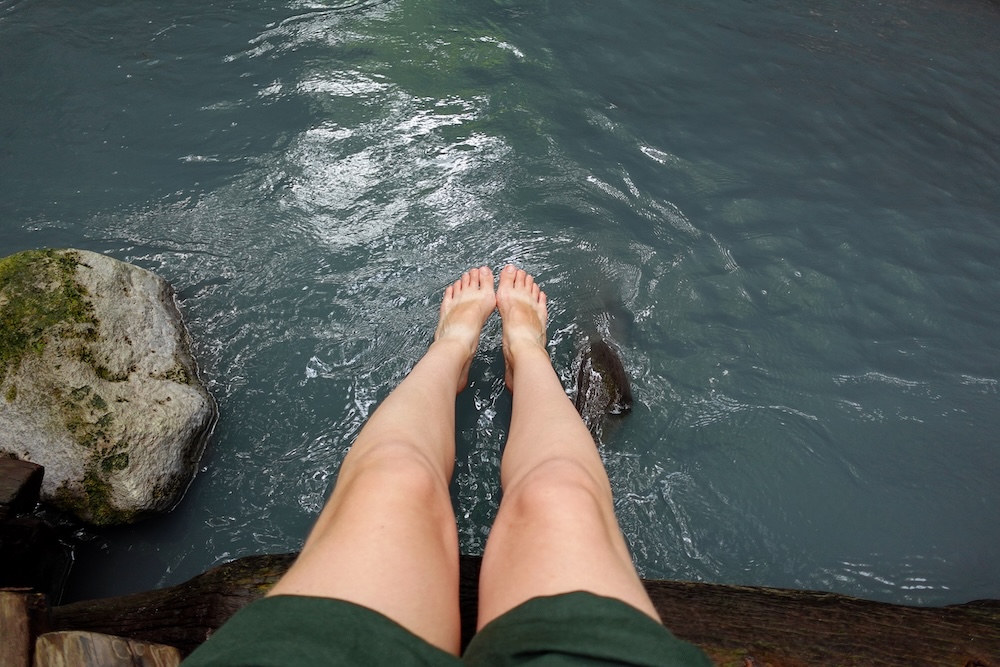 Feet Dipping Into the Milky Hot-Spring River at the Oyunuma River Natural Footbath, Noboribetsu Onsen, Japan – Audrey Bergner A first-person view of Audrey Bergner’s legs as she dips her feet into the milky blue-green water of the Oyunuma River Natural Footbath beside a mossy boulder in Noboribetsu Onsen, Hokkaido, Japan.