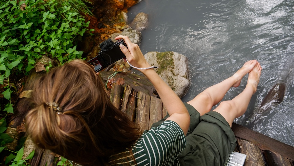 Feet in the Oyunuma River Natural Footbath on a wooden platform, Noboribetsu Onsen, Hokkaido, Japan – Audrey Bergner Seen from above, Audrey Bergner sits on a wooden platform with her bare feet in the flowing water of the Oyunuma River Natural Footbath, camera in hand, in the forest above Noboribetsu Onsen, Hokkaido, Japan.
