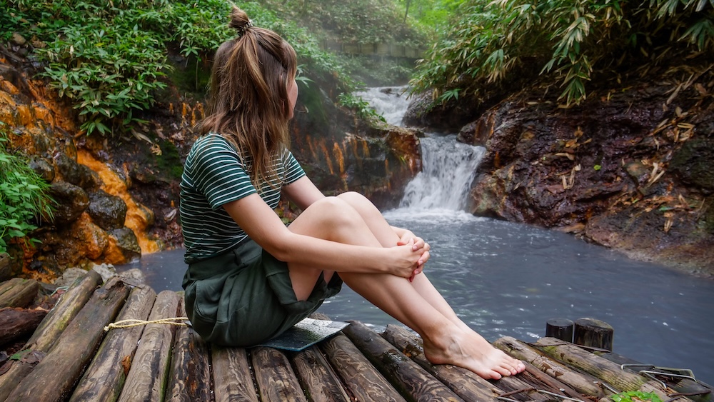 Audrey Bergner at Oyunuma River Natural Footbath in Noboribetsu Onsen, Hokkaido, Japan – Forest Waterfall Setting Audrey Bergner sits barefoot on a wooden platform beside the Oyunuma River Natural Footbath in Noboribetsu Onsen, Hokkaido, Japan, looking toward a small waterfall surrounded by dark volcanic rocks and lush green forest.