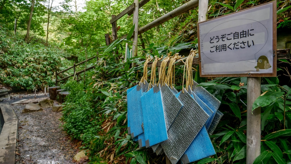 Free Sitting Mats for Visitors at Oyunuma River Natural Footbath, Noboribetsu Onsen, Hokkaido, Japan Blue and silver foam sitting mats hang from ropes beside a Japanese sign inviting visitors to use them freely at Oyunuma River Natural Footbath in Noboribetsu Onsen, Hokkaido, Japan, alongside a wooded footpath.