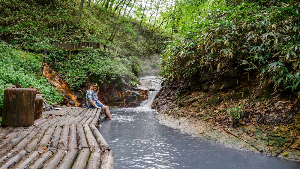 Samuel Jeffery sentado sobre una plataforma de troncos de madera con los pies en el agua geotermal lechosa del baño natural del río Oyunuma, en Noboribetsu Onsen, Hokkaido, Japón, rodeado de bosque, rocas teñidas de minerales y una pequeña cascada humeante.