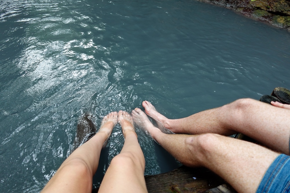Samuel Jeffery y Audrey Bergner remojando los pies en el río Oyunuma, Noboribetsu Onsen, Hokkaido, Japón Samuel Jeffery y Audrey Bergner remojan los pies descalzos en el agua geotérmica gris azulada del baño natural de pies del río Oyunuma, en Noboribetsu Onsen, Hokkaido, Japón, sentados sobre una plataforma de madera.