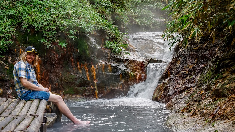 Cascada y asientos de bambú en el baño de pies del río Oyunuma, Noboribetsu Onsen, Japón – Samuel Jeffery Samuel Jeffery sentado descalzo sobre troncos de bambú con los pies en el agua termal lechosa del baño de pies natural del río Oyunuma, junto a una cascada humeante y rocas teñidas de naranja en Noboribetsu Onsen, Hokkaido, Japón.