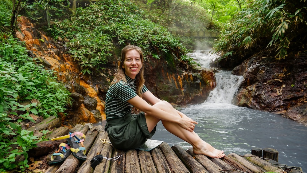 Riverside Log Seating, Forest and Steaming Cascade at the Oyunuma River Natural Footbath, Noboribetsu Onsen – Audrey Bergner Audrey Bergner smiles while sitting barefoot on log seating beside the Oyunuma River Natural Footbath, with her sandals next to her and a steaming cascade and lush forest behind, in Noboribetsu Onsen, Hokkaido, Japan.