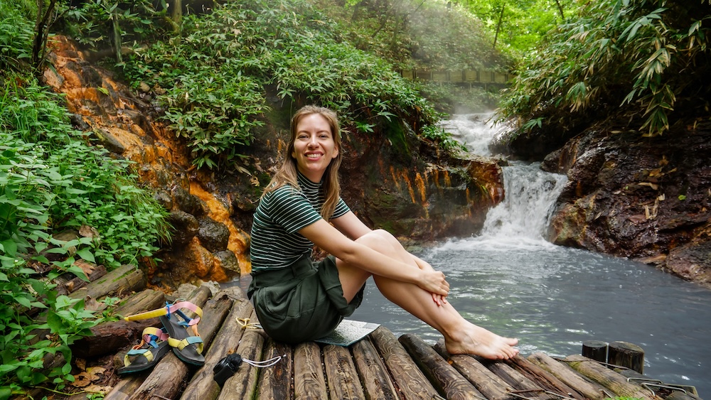 Asientos de tronco y cascada termal en el baño de pies natural del río Oyunuma, Noboribetsu Onsen, Hokkaido – Audrey Bergner Audrey Bergner sentada descalza sobre troncos junto a la cascada humeante del baño de pies natural del río Oyunuma, con sus sandalias al lado y roca teñida de naranja detrás, en Noboribetsu Onsen, Hokkaido, Japón.
